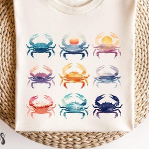 May include: Cream-colored t-shirt featuring a grid of nine watercolor crab designs in various colors. The crabs have sunset and ocean-themed patterns. The shirt is made of a soft, comfortable material.