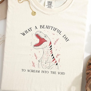 May include: Cream-colored Comfort Colors t-shirt with a dinosaur graphic. The dinosaur has a red mouth and black stripes. Text reads "WHAT A BEAUTIFUL DAY TO SCREAM INTO THE VOID." A gold necklace and sandals are visible.