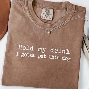 May include: Brown Comfort Colors t-shirt with the text "Hold my drink I gotta pet this dog" in white. A silver necklace with a pendant is on the shirt. The shirt is folded on a white surface.