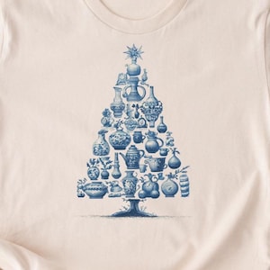 May include: A light beige t-shirt featuring a blue and white illustration of a Christmas tree made of various antique vases and teacups. The design is centered on the shirt.