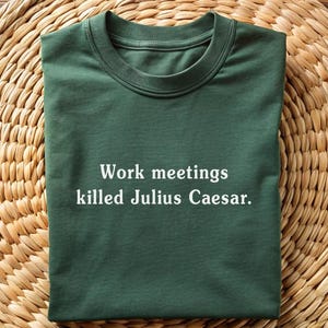 Könnte beinhalten: Ein dunkelgrünes T-Shirt mit weißem Text, der "Work meetings killed Julius Caesar." lautet.