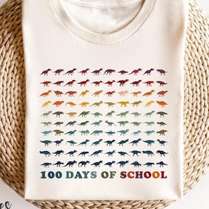 Puede incluir: Camiseta color crema con un diseño de dinosaurios colorido. El diseño incluye filas de siluetas de dinosaurios en un degradado arcoíris, con el texto "100 DAYS OF SCHOOL" en una combinación de colores a juego. La camiseta se muestra sobre una superficie tejida.