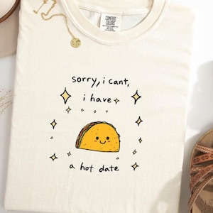 May include: Cream-colored t-shirt with the text "Sorry, I can't, I have a hot date" and a cartoon taco illustration. The taco has a smiling face and is surrounded by sparkling diamond shapes. The shirt has a Comfort Colors tag.