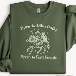 Könnte beinhalten: Olivgrünes Sweatshirt mit dem Text "Born to Dilly Dally Forced to Fight Fascists". Die Grafik zeigt einen Frosch, der mit einer Katze auf einem Pferd reitet. Das Sweatshirt hat lange Ärmel und einen Rundhalsausschnitt.