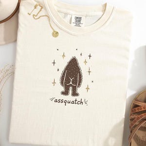 May include: Cream-colored t-shirt with a brown graphic of a Bigfoot-like creature, with the word "assquatch" below. The shirt has a crew neck and is surrounded by gold jewelry, a hat, and sandals.
