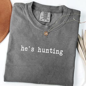 May include: A grey Comfort Colors t-shirt with the words "he's hunting" printed in white. A silver necklace with a small pendant lies on the shirt. The shirt is folded, and part of a brown hat is visible.