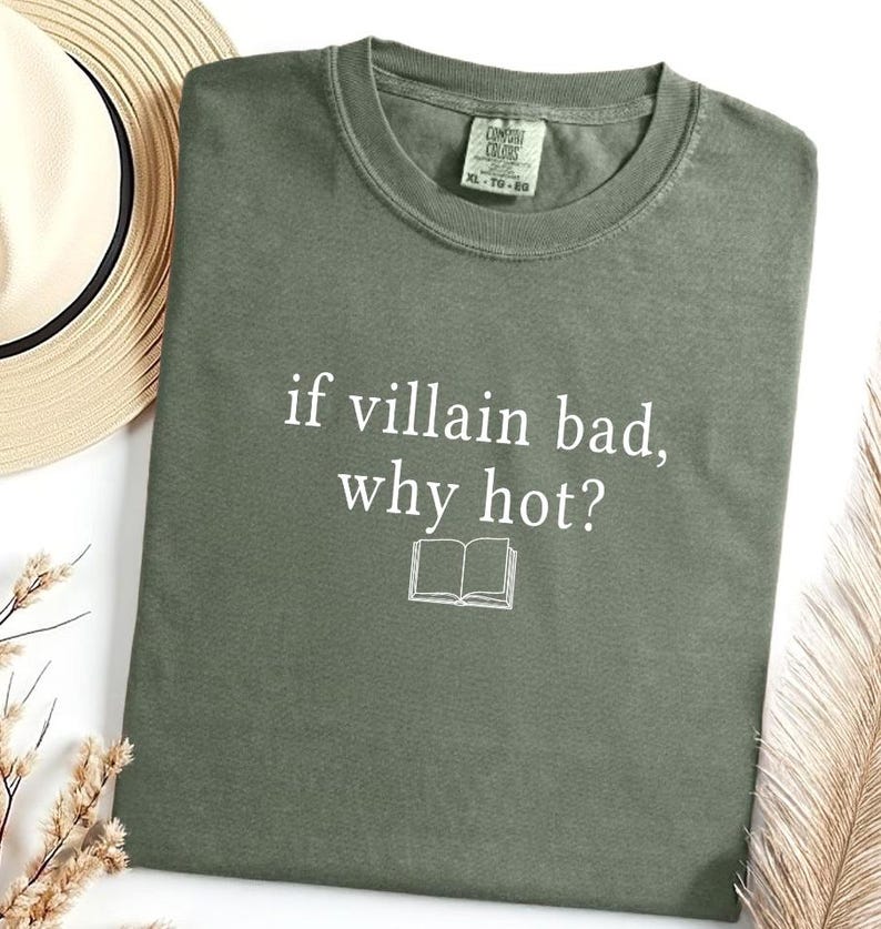 May include: An olive green t-shirt with the text "if villain bad, why hot?" in white, along with a small open book graphic. The shirt is a crew neck style.