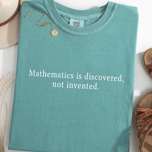 Pode incluir: Uma camiseta verde-azulado com o texto "Mathematics is discovered, not invented." em branco. Um colar dourado com um pingente redondo repousa sobre a camisa. A camisa está dobrada sobre uma superfície branca, com sandálias ao fundo.