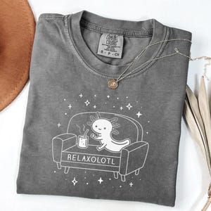 May include: A gray Comfort Colors t-shirt featuring a white graphic of an axolotl relaxing on a couch with a cup of coffee. The text "RELAXOLOTL" is below the couch. A silver necklace with a pendant is on the shirt.