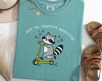 May include: Teal-colored t-shirt with a raccoon riding a yellow scooter, and the text "Off to improve mobility." The shirt also has the text "customize! booootees."