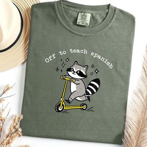 Op de afbeelding: Olijfgroen t-shirt met de tekst "Off to teach spanish" en een cartoon wasbeer op een gele scooter. De wasbeer is grijs, wit en zwart. Het shirt is van het merk Comfort Colors.
