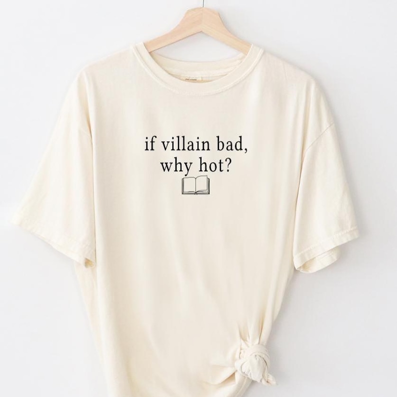 May include: Cream-colored t-shirt with the text "if villain bad, why hot?" in black, along with a small book illustration. The shirt is hanging on a wooden hanger against a white background. The t-shirt has a relaxed fit.