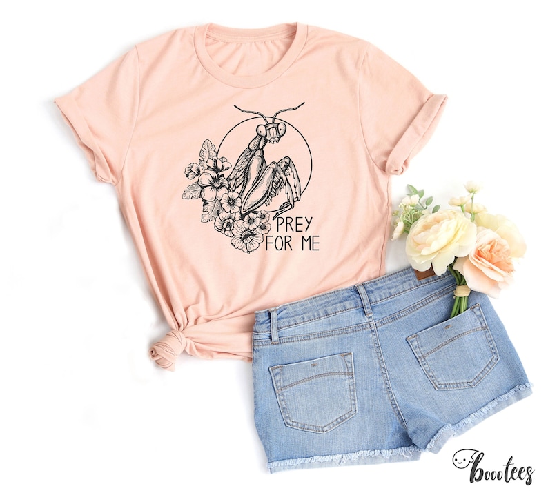May include: A peach-colored t-shirt with a black and white illustration of a praying mantis with flowers and the text "Prey for Me".
