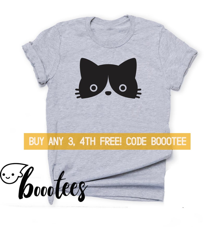 May include: A grey t-shirt with a black and white cat face graphic. The cat's face is centrally placed, with large eyes and a small nose. A yellow banner displays the text "BUY ANY 3, 4TH FREE! CODE BOOOTEE".