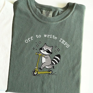 Könnte beinhalten: Ein gefaltetes, salbeigrünes T-Shirt mit dem Text "Off to write IEPs" und einem Cartoon-Waschbären auf einem gelben Roller. Das Shirt hat ein Comfort Colors-Etikett am Ausschnitt.