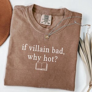 May include: A brown Comfort Colors t-shirt with the text "if villain bad, why hot?" and a small book graphic. A silver necklace with a pendant rests on the shirt. The shirt is size S-P-CH.