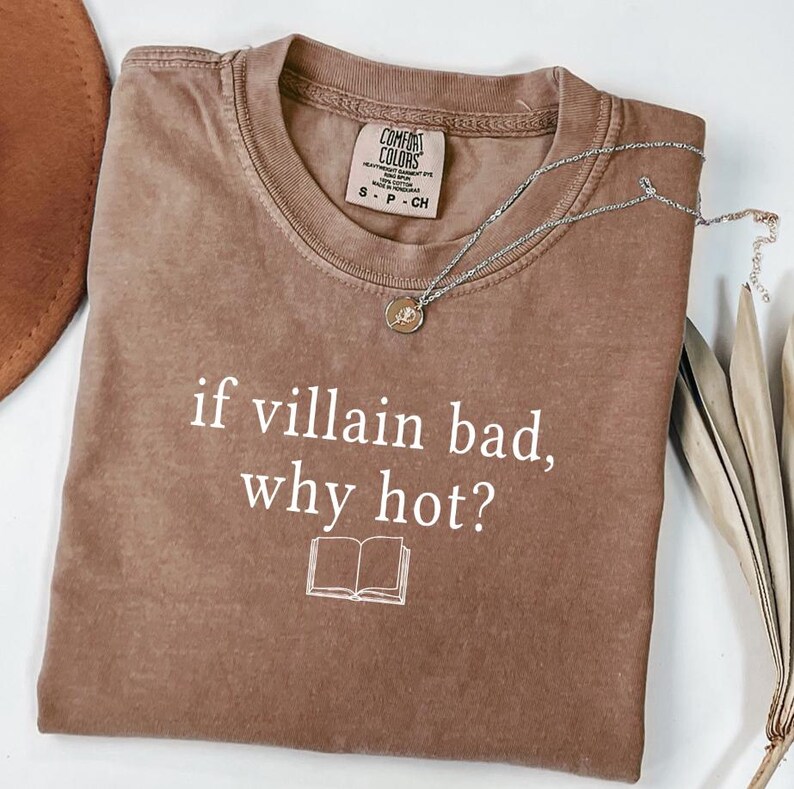 May include: A brown Comfort Colors t-shirt with the text "if villain bad, why hot?" and a small book graphic. A silver necklace with a pendant rests on the shirt. The shirt is size S-P-CH.