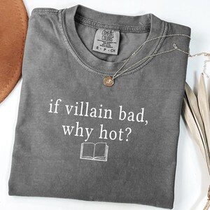 May include: A gray t-shirt with the text "if villain bad, why hot?" in white, along with a small book graphic. A silver necklace with a round pendant is draped across the shirt. The shirt is folded neatly.