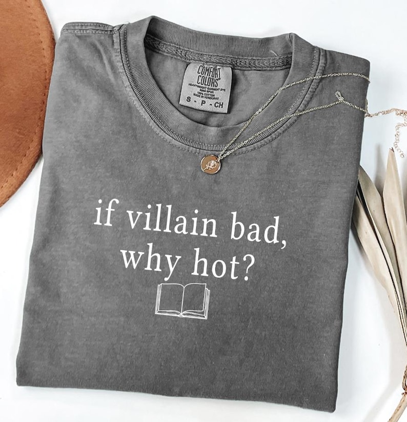May include: A gray t-shirt with the text "if villain bad, why hot?" in white, along with a small book graphic. A silver necklace with a round pendant is draped across the shirt. The shirt is folded neatly.