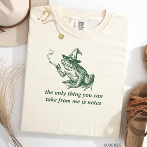 May include: A cream-coloured t-shirt featuring a green illustration of a frog in a wizard's hat, smoking a cigarette. The text "the only thing you can take from me is notes" is printed below. This is a clothing item.