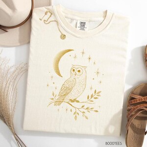 Könnte beinhalten: Cremefarbenes Comfort Colors Sweatshirt mit einer goldenen Illustration einer Eule auf einem Ast, einem Mond und Sternen. Der Text "BOOOTEES" ist unten rechts aufgedruckt.