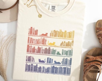 May include: Cream-coloured t-shirt with a colourful bookshelf design and cats. The graphic shows rows of books in a rainbow of colours, with a cat silhouette on each shelf. The shirt has a crew neck and short sleeves. The brand name "COMFORT COLORS" is printed on the shirt.