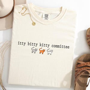 Op de afbeelding: Crèmekleurig T-shirt met de zwarte tekst "itty bitty kitty committee". Daaronder drie cartoonkatten in grijs, oranje en grijs. Een gouden ketting en sandalen zijn ook zichtbaar.