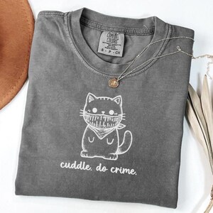 May include: A gray Comfort Colors t-shirt with a white graphic of a cat wearing a bandana. The text "cuddle. do crime." is below the cat. A silver necklace with a small pendant is on the shirt.