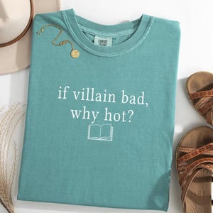 May include: A teal-colored t-shirt with the text "if villain bad, why hot?" and a book graphic. A gold necklace and a beige hat are visible. Brown sandals are placed to the right of the shirt.