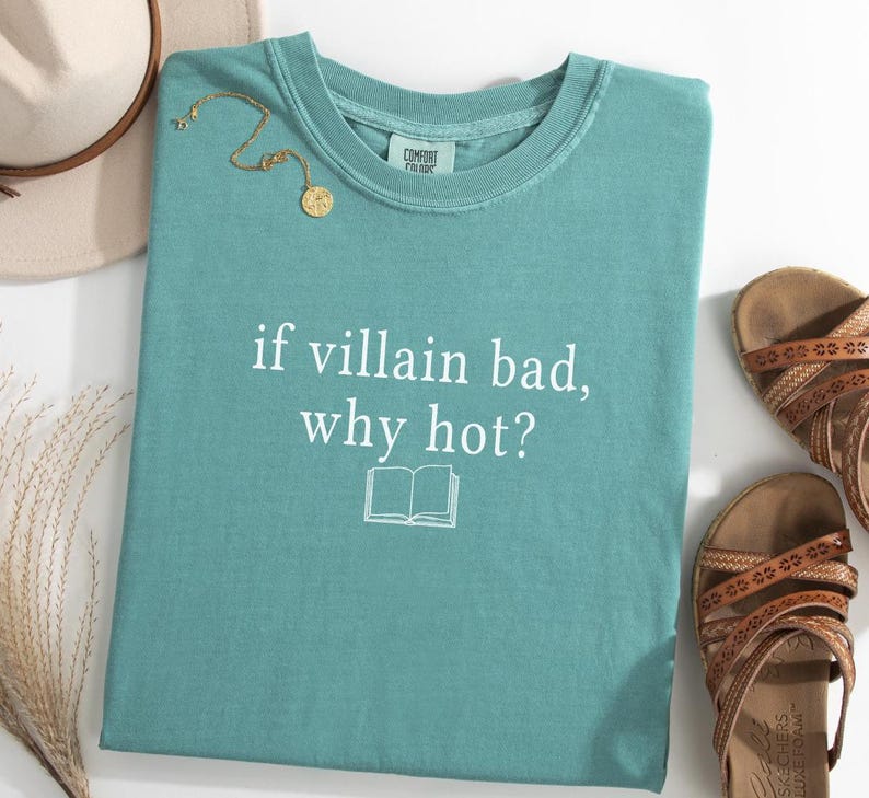 May include: A teal-colored t-shirt with the text "if villain bad, why hot?" and a book graphic. A gold necklace and a beige hat are visible. Brown sandals are placed to the right of the shirt.