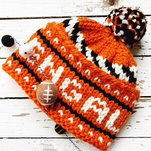 May include: An orange, black, and white knitted beanie hat with a pom-pom. The hat features a repeating pattern and a small felt football attached. Decorative felt balls in black, white, and orange are strung nearby.
