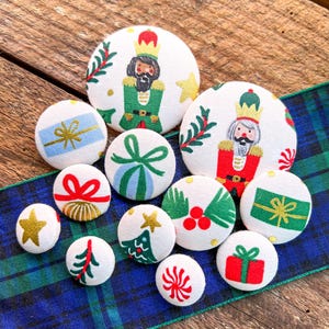 May include: A collection of white fabric-covered buttons with Christmas-themed designs. The buttons feature images of nutcrackers, gifts, bows, Christmas trees, and stars. They are arranged on a blue and green plaid ribbon and a wooden surface.