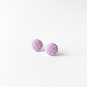 May include: Two circular fabric-covered buttons featuring a small-scale checkered pattern in light pink and pale blue. The buttons have a rounded, domed shape and a textured textile surface, presented as a pair of matching round fasteners.
