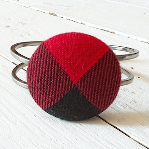 May include: A silver bangle bracelet with a red and black plaid fabric button. The button is round and has a geometric pattern.
