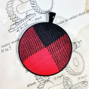 May include: A round pendant made from black and red plaid fabric with a black metal bail. The pendant has a glossy finish.