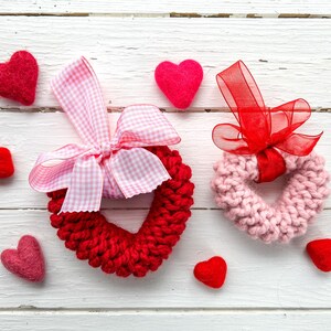 Può includere: Due ghirlande di cuori all'uncinetto, una rossa e una rosa, con fiocchi di nastro rosa e rosso. Le ghirlande sono su uno sfondo di legno bianco con piccoli cuori di feltro sparsi intorno.