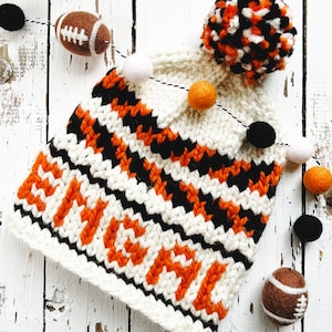 May include: A cream-coloured knitted hat with orange and black chevron stripes and the word "BENGAL" in orange. The hat has a large pom-pom and is decorated with felt American footballs and black, white, and orange pom-poms.
