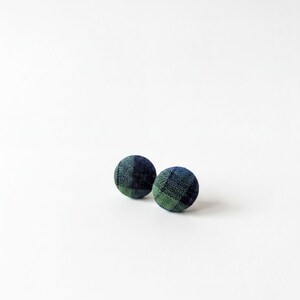 May include: Two circular fabric-covered buttons featuring a dark navy blue and forest green plaid pattern. The buttons have a rounded, dome-like shape with a visible woven textile texture throughout the surface of the fabric covering each button.
