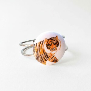 May include: A silver-toned metal cuff bracelet featuring a circular fabric-covered centerpiece. The fabric displays a detailed illustration of an orange and black tiger with white accents, rendered in a sketch-like style on a light background.