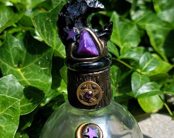 May include: A clear glass bottle with a purple and black stone stopper. The stopper features a crescent moon and a star. The bottle has a purple and black stone charm on the front.
