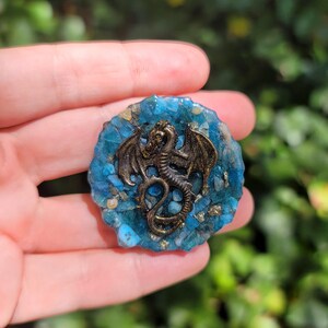 May include: A round, blue and gold resin pendant with a bronze dragon centerpiece. The dragon has outstretched wings and intricate details. The pendant is approximately 2 inches in diameter, with a textured, stone-like appearance.