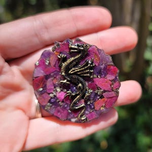 May include: A round, handcrafted pendant featuring a bronze dragon over a mosaic of purple and pink gemstones. The dragon is intricately detailed, and the gemstones are set in a resin base. A unique, artistic piece.