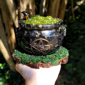 May include: A black resin cauldron with gold accents and a gold pentagram, filled with green bubbling liquid. The cauldron sits on a moss-covered wooden base, evoking a mystical theme.