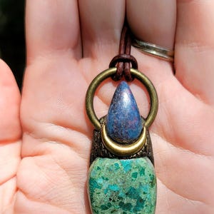 May include: A handcrafted pendant featuring a teardrop-shaped green stone with blue and teal inclusions, topped with a blue stone and a brass ring. The pendant hangs from a brown leather cord.