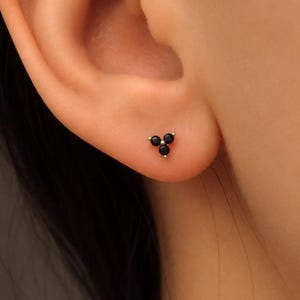 Könnte beinhalten: Ein kleiner Ohrstecker mit drei winzigen, runden, schwarzen Steinen, die in einer dreieckigen Gruppe angeordnet sind. Die Steine sind in einer metallischen Basis gefasst und bilden ein kompaktes, dreizackiges Design auf dem Ohrläppchen.