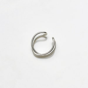 May include: A silver-colored ear cuff with a minimalist design. The cuff features two intertwined, curved bands that create a crossed effect. The jewelry is set against a white background.