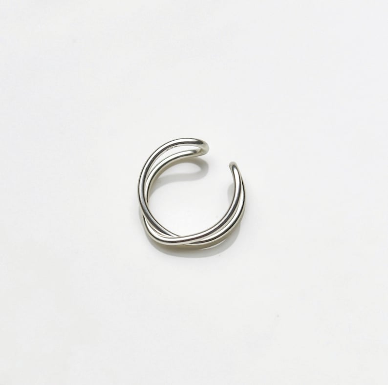 May include: A silver-colored ear cuff with a minimalist design. The cuff features two intertwined, curved bands that create a crossed effect. The jewelry is set against a white background.