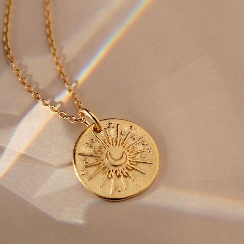 May include: A gold-toned necklace featuring a circular pendant with an embossed sun and crescent moon design surrounded by small stars. The pendant hangs from a delicate gold-toned chain, catching light to create a subtle rainbow prism effect.