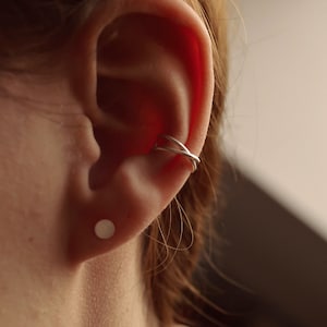 May include: Close-up of an ear with a silver ear cuff and a small, round stud earring. The ear cuff has a crisscross design. The earring is a simple, flat circle. The skin tone is fair, and the background is a soft, neutral color.