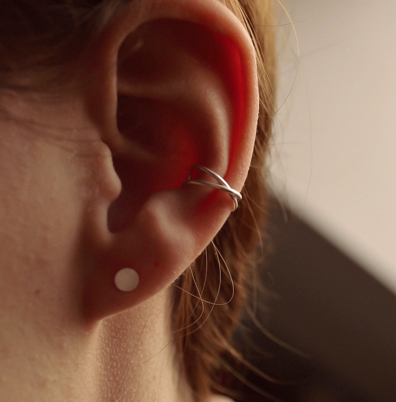 May include: Close-up of an ear with a silver ear cuff and a small, round stud earring. The ear cuff has a crisscross design. The earring is a simple, flat circle. The skin tone is fair, and the background is a soft, neutral color.
